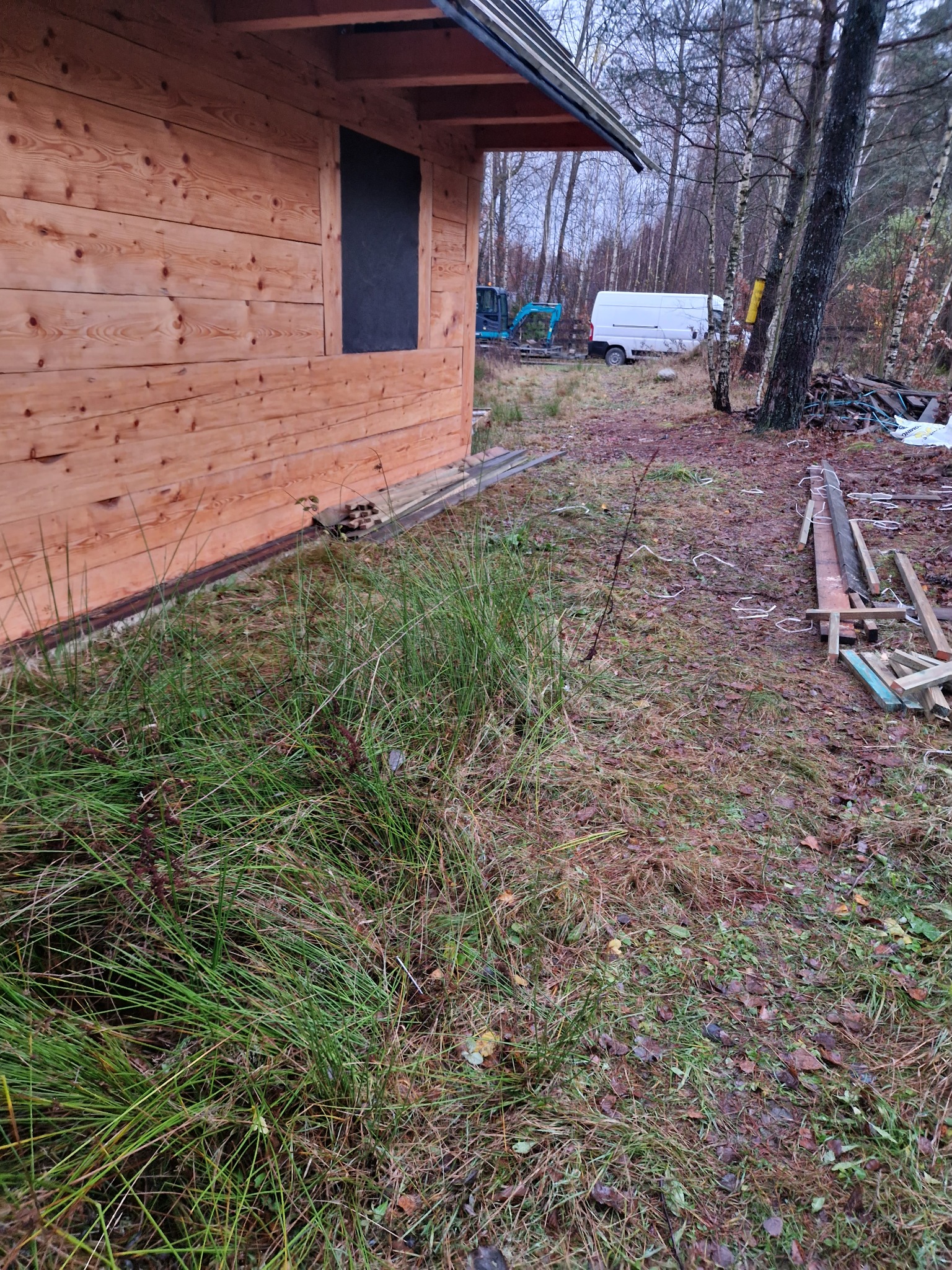 Fragment drewnianej konstrukcji budynku w stanie surowym, z widoczną deską obiciową i otworem okiennym, na tle zarośniętego terenu z widoczną koparką i samochodem dostawczym w oddali.