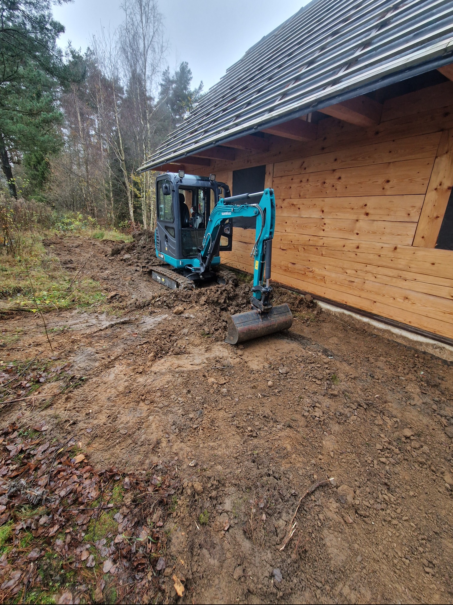 Niebieska mini koparka wykonuje prace ziemne przy drewnianym budynku w jesiennej scenerii, widoczne świeżo przekopane błoto i drzewa w tle.