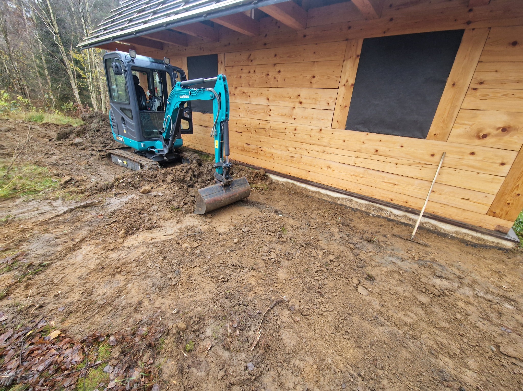 Niebieska mini koparka Sunward 20P wykonuje wykop przy drewnianym budynku z zabezpieczonymi oknami, widoczne ślady opon i świeżo naruszona ziemia.