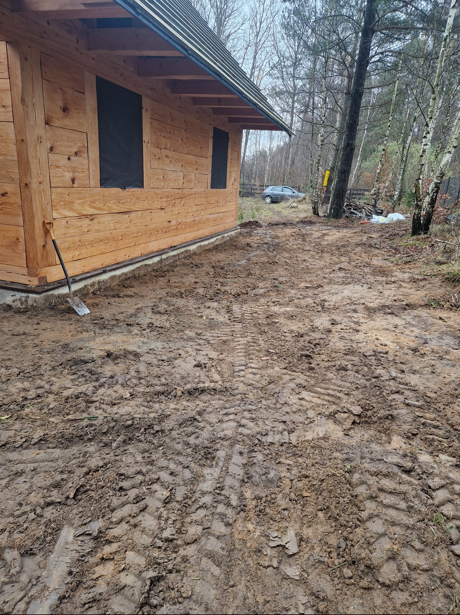 Świeżo wybudowany drewniany domek z czarnymi zaślepkami w oknach, z ziemią wyrównaną wokół fundamentów i śladami opon, łopata oparta o ścianę budynku.