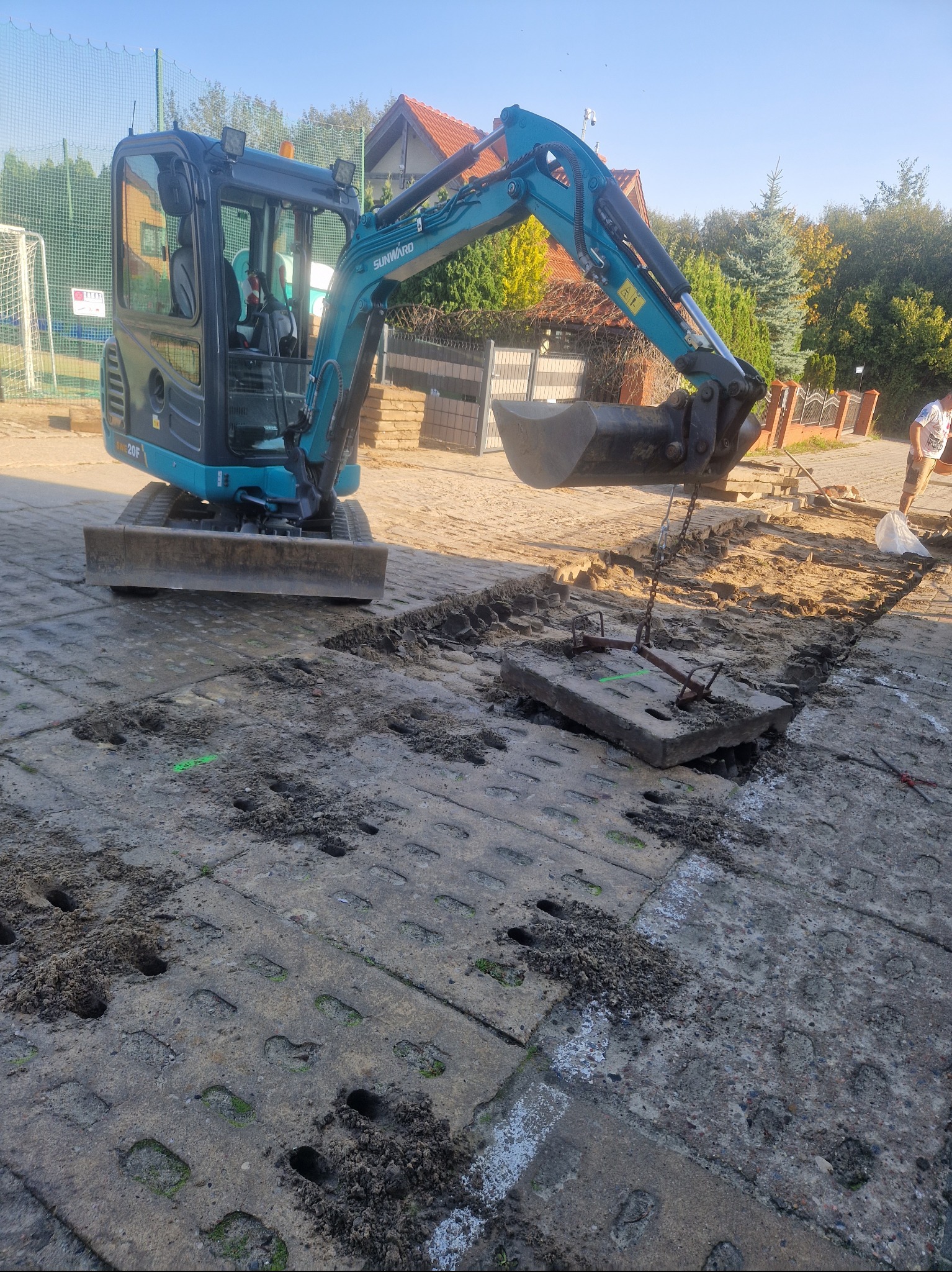 Niebieska mini koparka Sunward SW20F podnosi betonową płytę z ziemi za pomocą łańcuchów, widoczne ślady wykopu w kostce brukowej, w tle ogrodzenie i dom.