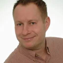 Uśmiechnięty mężczyzna w pomarańczowej koszuli w kratę, z krótkimi brązowymi włosami, na jasnym tle. Portret z lekkim przekrzywieniem głowy.