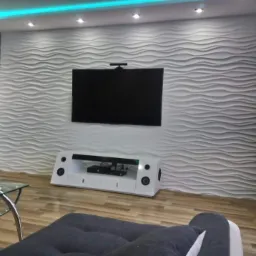 Nowoczesny salon z białą ścianą 3D o falistym wzorze, podświetleniem LED na suficie i telewizorem LCD zawieszonym nad białą szafką RTV z głośnikami.