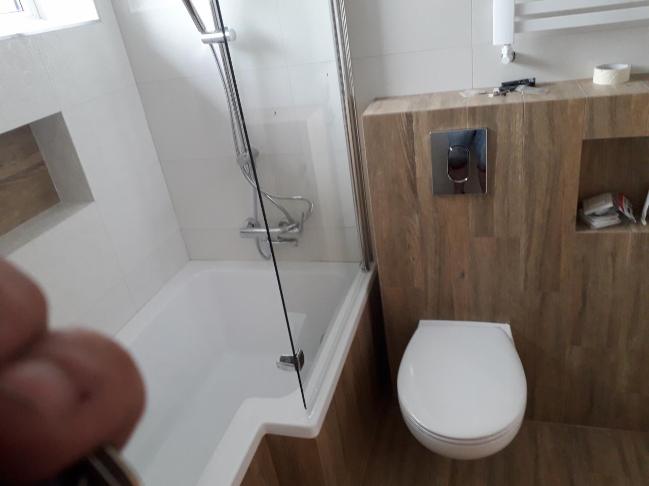 Nowoczesna łazienka po remoncie: biała wanna z parawanem nawannowym, toaleta z chromowanym przyciskiem spłukiwania, drewnopodobne płytki na ścianach i podłodze, wnęka w ścianie z akcesoriami...