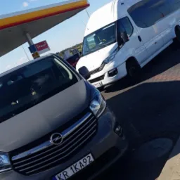 Szary samochód osobowy i biały bus zaparkowane przy stacji benzynowej Shell w słoneczny dzień.