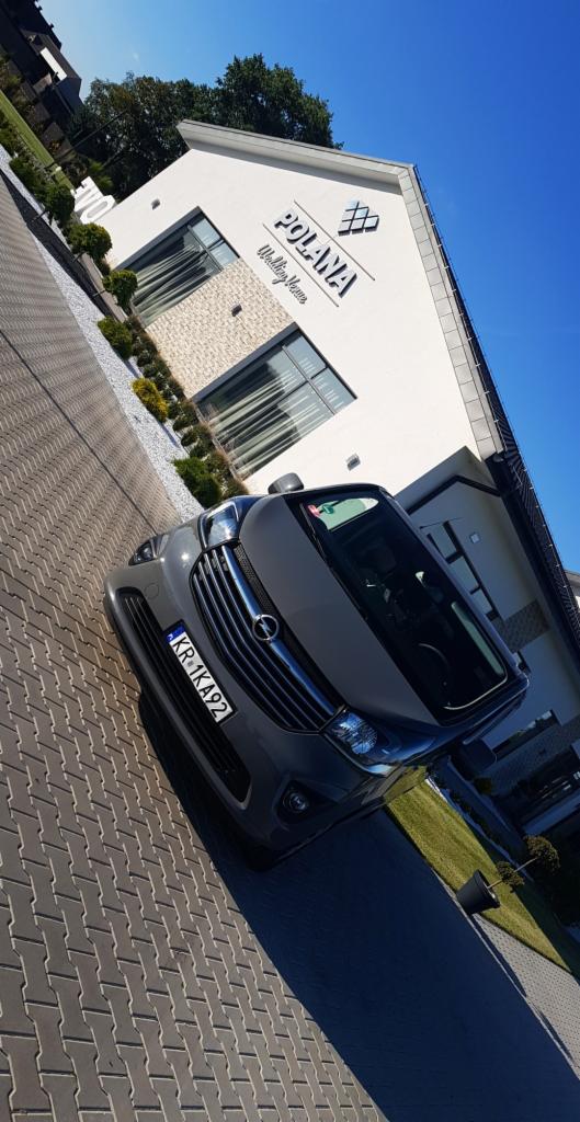 Szary bus marki Opel Vivaro z numerem rejestracyjnym KR 1KA92 zaparkowany przed budynkiem weselnym 'Polana Wedding Venue' w słoneczny dzień.