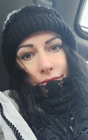 Portret kobiety w czapce i szaliku, z delikatnym uśmiechem, uchwycony w naturalnym świetle z bliska.