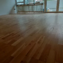 Położona jasna podłoga drewniana w pustym pokoju z grzejnikiem i balkonem. Widoczne detale usłojenia drewna. Naturalne światło.