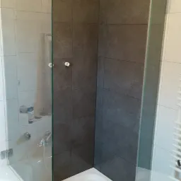 Wykończona kabina prysznicowa z grafitowymi płytkami na ścianach i szklanymi drzwiami. Widoczna biała brodzik i fragment wanny. Oświetlenie górne, neutralne.