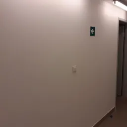 Biała ściana korytarza z zamontowanym znakiem ewakuacyjnym i włącznikiem światła, widoczny fragment podłogi z brązowych płytek i oświetlenie sufitowe.