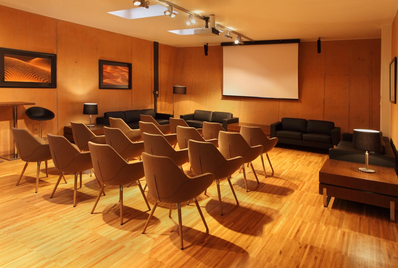 Sala konferencyjna z rzędami beżowych krzeseł, czarnymi sofami, projektorem i ekranem projekcyjnym na pomarańczowych ścianach, oświetlona punktowymi lampami i lampami stołowymi.