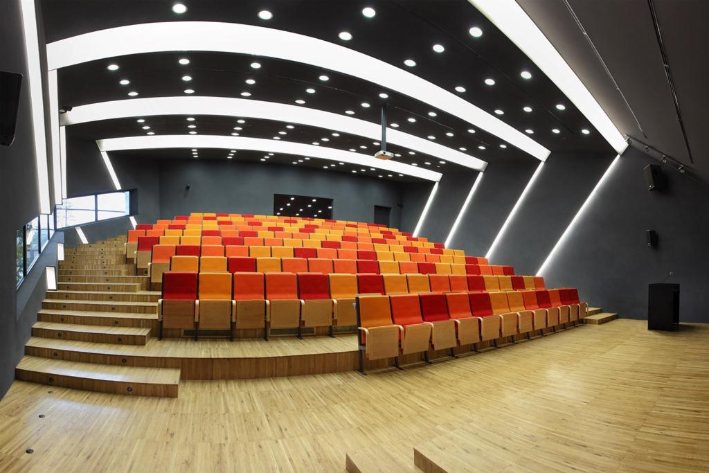 Widok z przodu na pustą salę konferencyjną z rzędami foteli w odcieniach czerwieni i pomarańczy, oświetloną nowoczesnymi lampami sufitowymi i punktowymi, z drewnianą podłogą i schodami.