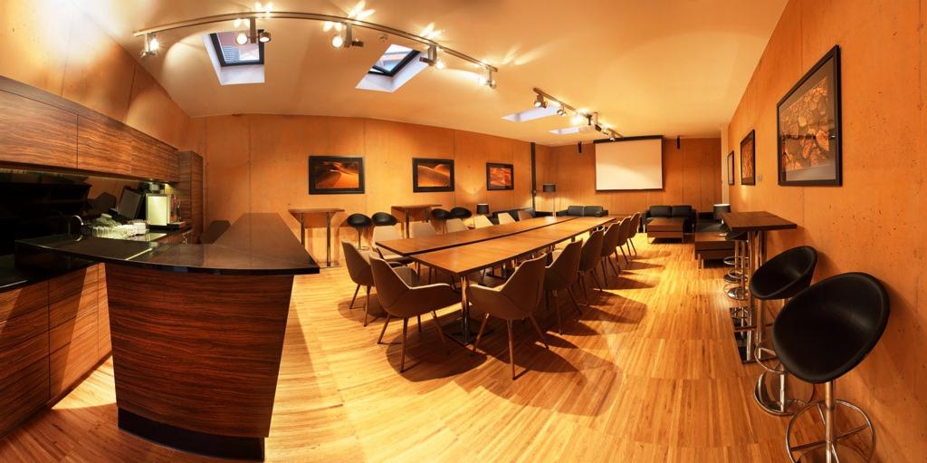 Sala konferencyjna z drewnianą podłogą, długim stołem z krzesłami, barkiem z ciemnym blatem i projektorem z ekranem w tle.