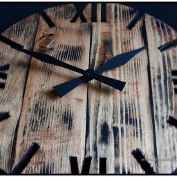 Krausaky Krzysztof Krause - Drewniany zegar ścienny w stylu rustykalnym z czarnymi wskazówkami i rzymskimi cyframi wyciętymi w drewnie, widoczna faktura drewna z opalonymi elementami.