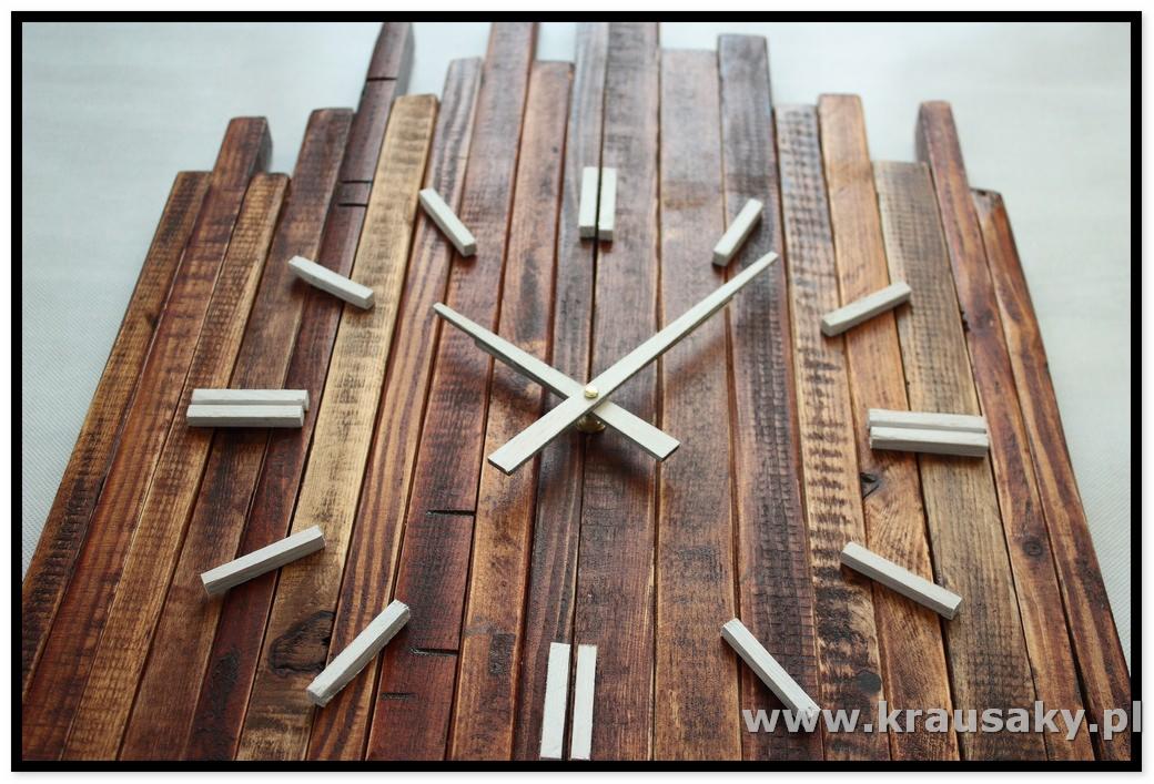 Unikatowy zegar ścienny wykonany z postarzanych desek o zróżnicowanej wysokości, z jasnymi wskazówkami i oznaczeniami godzin w formie drewnianych klocków.