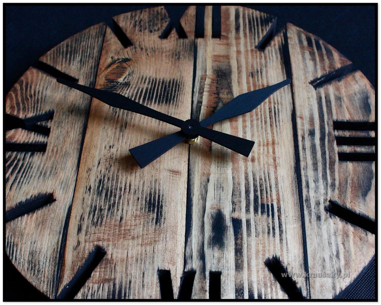 Drewniany zegar ścienny w stylu rustykalnym z czarnymi wskazówkami i rzymskimi cyframi wyciętymi w drewnie, widoczna faktura drewna z opalonymi elementami.