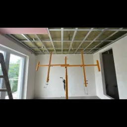 Montaż metalowej konstrukcji sufitu podwieszanego z izolacją wełną mineralną, widoczny podnośnik do płyt, okno i fragment drabiny.