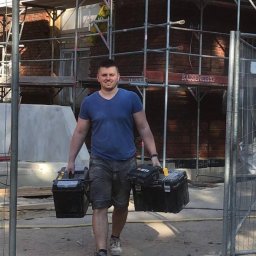 Tron - Pracownik budowlany niosący dwie skrzynki narzędziowe na tle rusztowania przy ceglanym budynku w trakcie remontu.