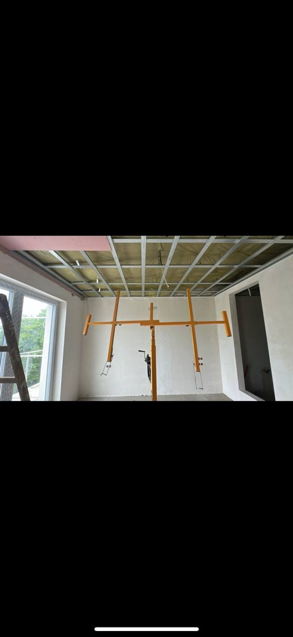 Montaż metalowej konstrukcji sufitu podwieszanego z izolacją wełną mineralną, widoczny podnośnik do płyt, okno i fragment drabiny.