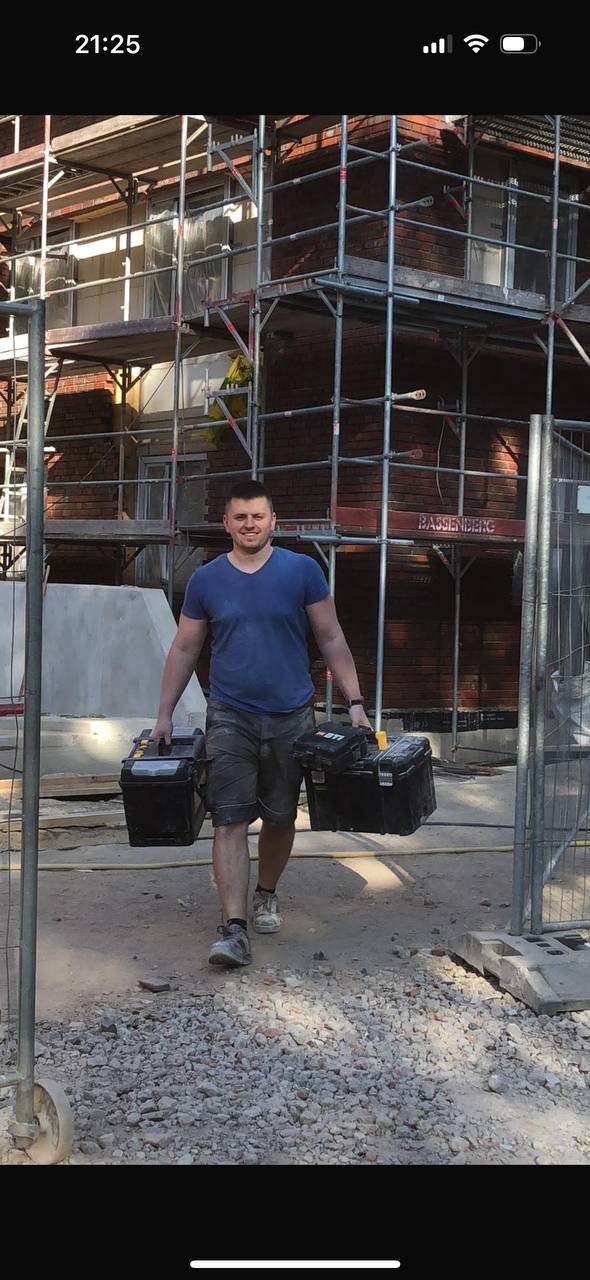 Pracownik budowlany niosący dwie skrzynki narzędziowe na tle rusztowania przy ceglanym budynku w trakcie remontu.