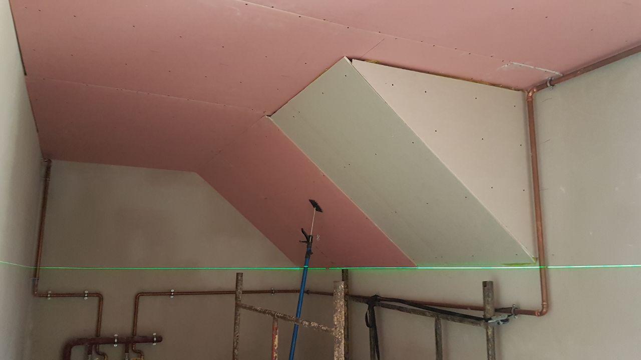 Montaż płyt kartonowo-gipsowych na skośnym poddaszu, widoczna konstrukcja metalowa, rury miedziane i pozioma linia lasera.