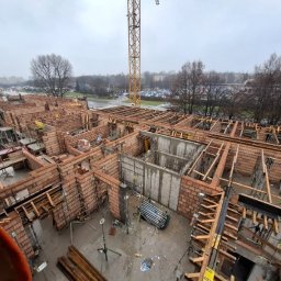 Good-Bud Kamil Stachura - Wysoki kadr z góry przedstawiający plac budowy z wzniesionymi ceglanymi ścianami i betonowymi konstrukcjami, widocznym drewnianym szalunkiem i żółtym dźwigiem. Mokry teren.