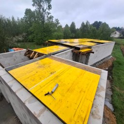 Good-Bud Kamil Stachura - Konstrukcja ścian parteru z szarych bloczków, z przygotowanym żółtym deskowaniem pod strop i pistoletem do uszczelniania na pierwszym planie, w tle zielone drzewa.