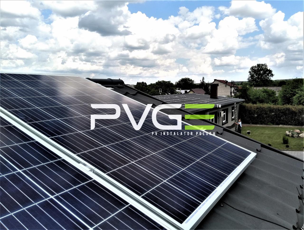 Panele słoneczne na dachu domu jednorodzinnego z widocznym logo PVG i napisem PV INSTALATOR POLSKA, w tle błękitne niebo z chmurami i zieleń trawnika.