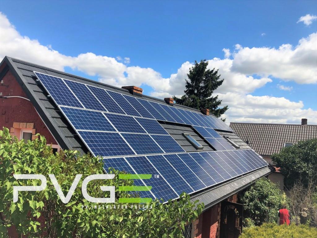 Instalacja paneli słonecznych na dachu domu jednorodzinnego z oknami dachowymi, widok z poziomu ogrodu, błękitne niebo z chmurami w tle.