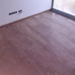 Ułożona brązowa podłoga z płytek imitujących kamień w pomieszczeniu w Sosnowcu, widoczne gniazdka elektryczne na ścianie i okno zabezpieczone folią ochronną.