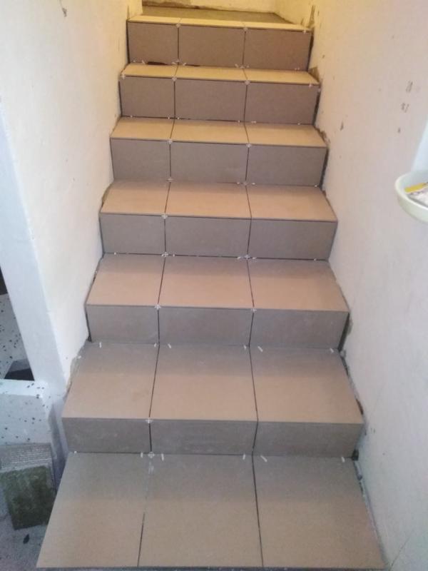 Schody w trakcie układania płytek ceramicznych w odcieniach beżu, widoczne krzyżyki dystansowe i niedokończone fugi.