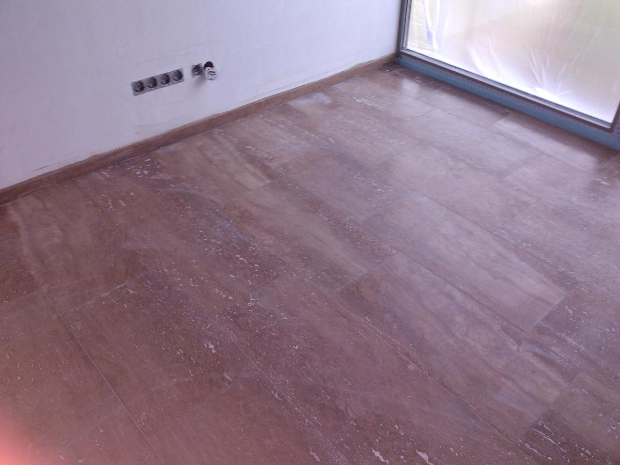 Ułożona brązowa podłoga z płytek imitujących kamień w pomieszczeniu w Sosnowcu, widoczne gniazdka elektryczne na ścianie i okno zabezpieczone folią ochronną.