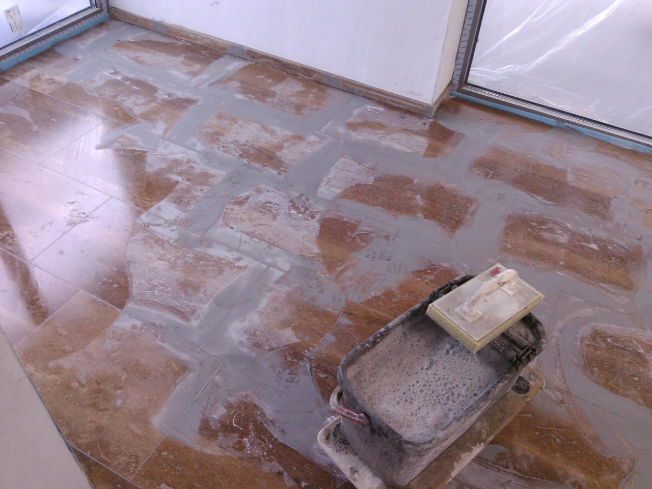 Brązowe płytki podłogowe z fugą w trakcie nakładania, wiadro z wodą i gąbką w rogu pokoju.