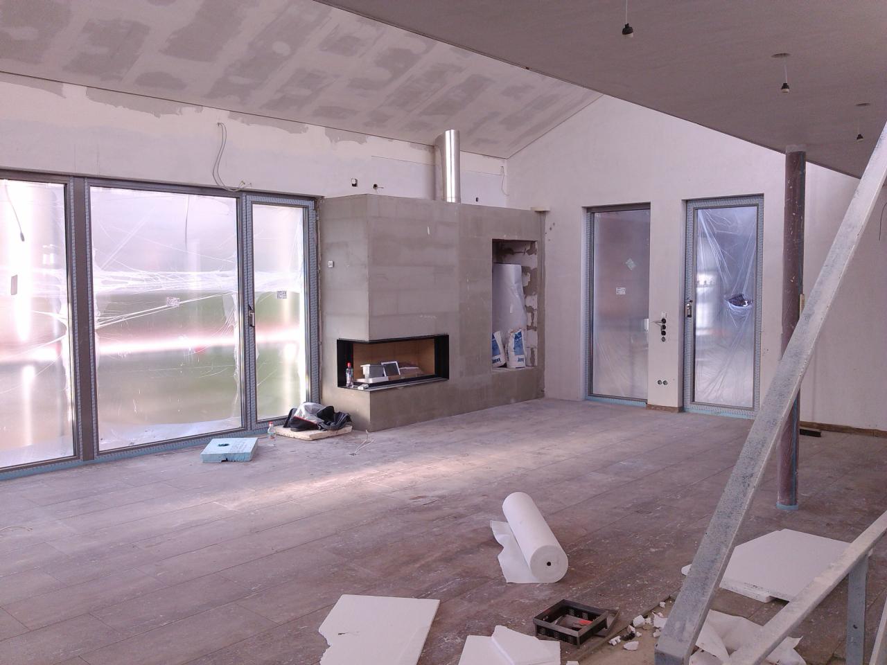 Wnętrze w trakcie remontu: surowe ściany, widoczne szpachlowanie sufitu, kominek obłożony surowym betonem, osłonięte folią drzwi, panele podłogowe, rozłożone materiały budowlane i drabina.