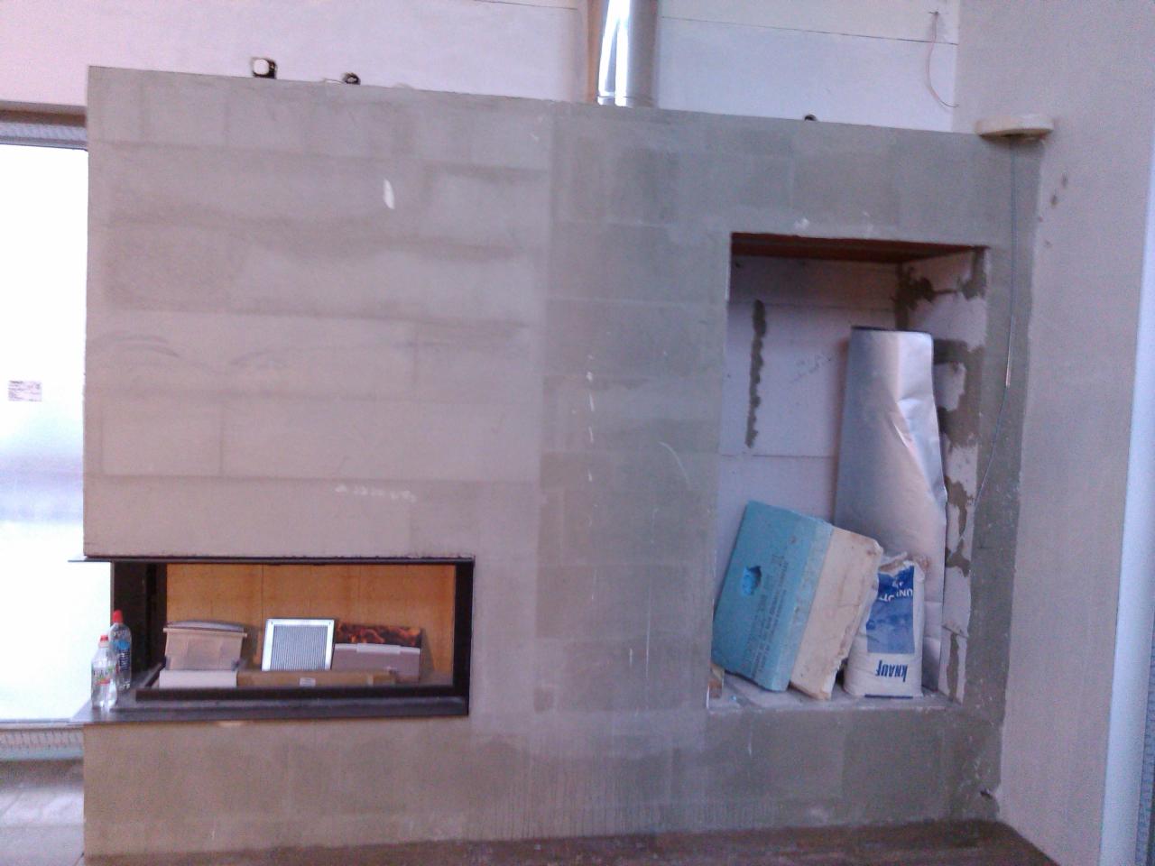 Wnętrze w trakcie budowy kominka z widoczną wkładką kominkową, obudową z bloczków i wnęką z materiałami budowlanymi.