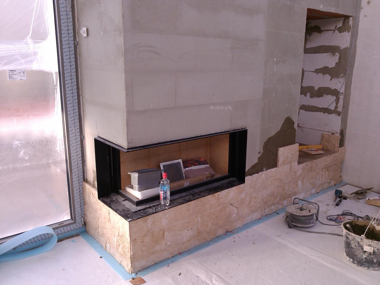 Wnętrze w trakcie wykańczania kominka z obudową z trawertynu, widoczne narzędzia i materiały budowlane, okno zabezpieczone folią malarską.