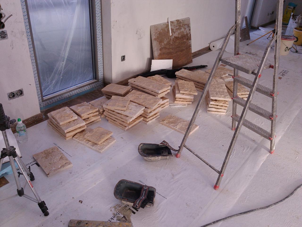 Przygotowane do ułożenia płytki podłogowe z naturalnego kamienia, ułożone w stosy na folii ochronnej, obok drabina aluminiowa i poziomica, w tle okno i fragment ściany w trakcie remontu.