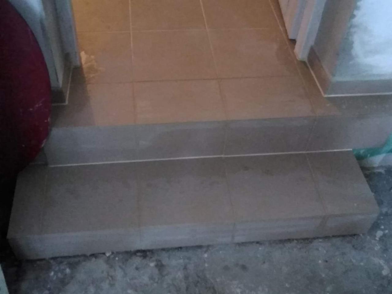 Wykończone schody wejściowe do pomieszczenia, pokryte jasnymi, kwadratowymi płytkami ceramicznymi, z widocznymi fugami i fragmentem surowego podłoża.