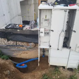 Otwarta szafka elektryczna z licznikiem i zabezpieczeniami, częściowo zamontowana na zewnątrz budynku, z widocznymi kablami i narzędziami, oraz wykopanym dołem z niebieskim przewodem karbowanym.