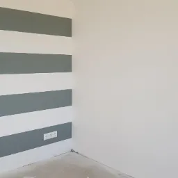 Pomieszczenie po malowaniu ścian w poziome pasy szaro-białe, widoczne włączniki światła i gniazdka elektryczne.