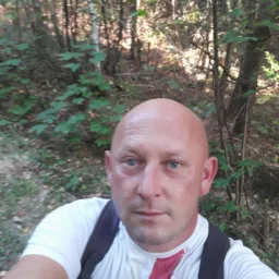 Portret mężczyzny z plecakiem na tle leśnym, z widoczną częścią białej koszulki z czerwonym wzorem. Oświetlenie naturalne, kadr z bliska.