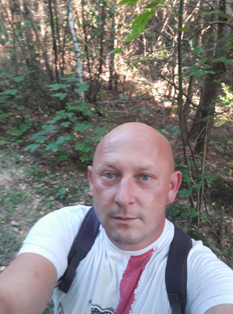 Portret mężczyzny z plecakiem na tle leśnym, z widoczną częścią białej koszulki z czerwonym wzorem. Oświetlenie naturalne, kadr z bliska.