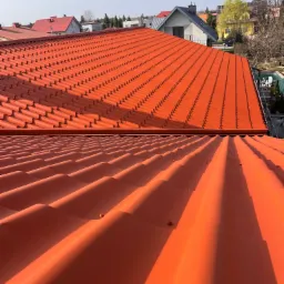 Dwa dachy z czerwonej blachodachówki, jeden falisty, drugi z drobnymi poprzecznymi przetłoczeniami, w tle zabudowa jednorodzinna pod błękitnym niebem.