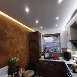 Nowoczesna kuchnia z dekoracyjną ścianą w odcieniach brązu, oświetleniem LED podsufitowym i drewnianymi szafkami, widok na zlew z naczyniami, ekspres do kawy i gotujący się garnek na płycie...