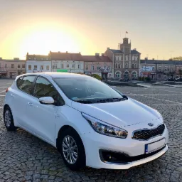 Biały samochód Kia Ceed na brukowanym placu miejskim o wschodzie słońca, z budynkami w tle i fontanną.