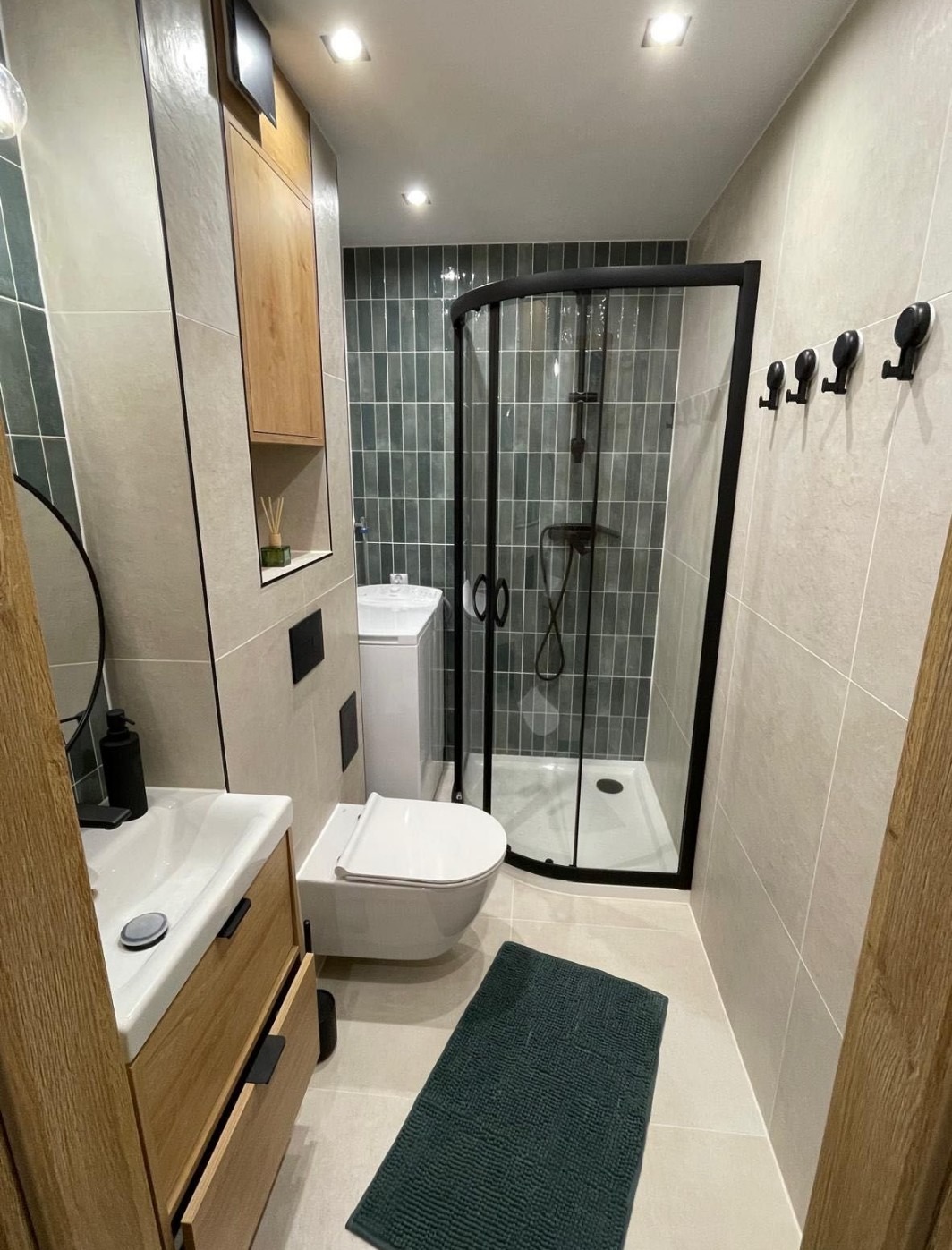 Nowoczesna łazienka z kabiną prysznicową typu walk-in, umywalką z szafką z drewna i wiszącą toaletą. Szaro-zielone płytki na ścianach i podłodze. Minimalistyczny design.