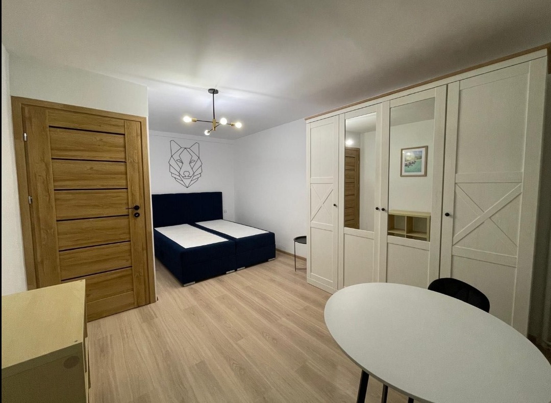 Jasne wnętrze pokoju z drewnianą podłogą, białymi ścianami, łóżkiem z granatowym wezgłowiem, dużą szafą z lustrami i drewnianymi drzwiami. Na ścianie grafika wilka.