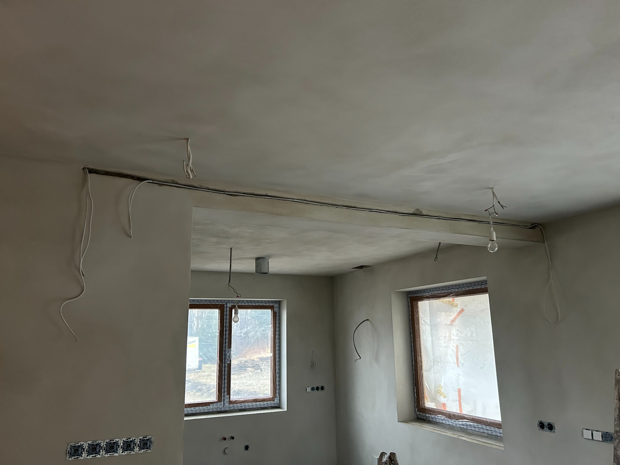 Wnętrze w trakcie prac wykończeniowych: gładzie na ścianach i suficie, widoczne kable elektryczne, okna zabezpieczone folią. Przygotowanie do dalszych prac.