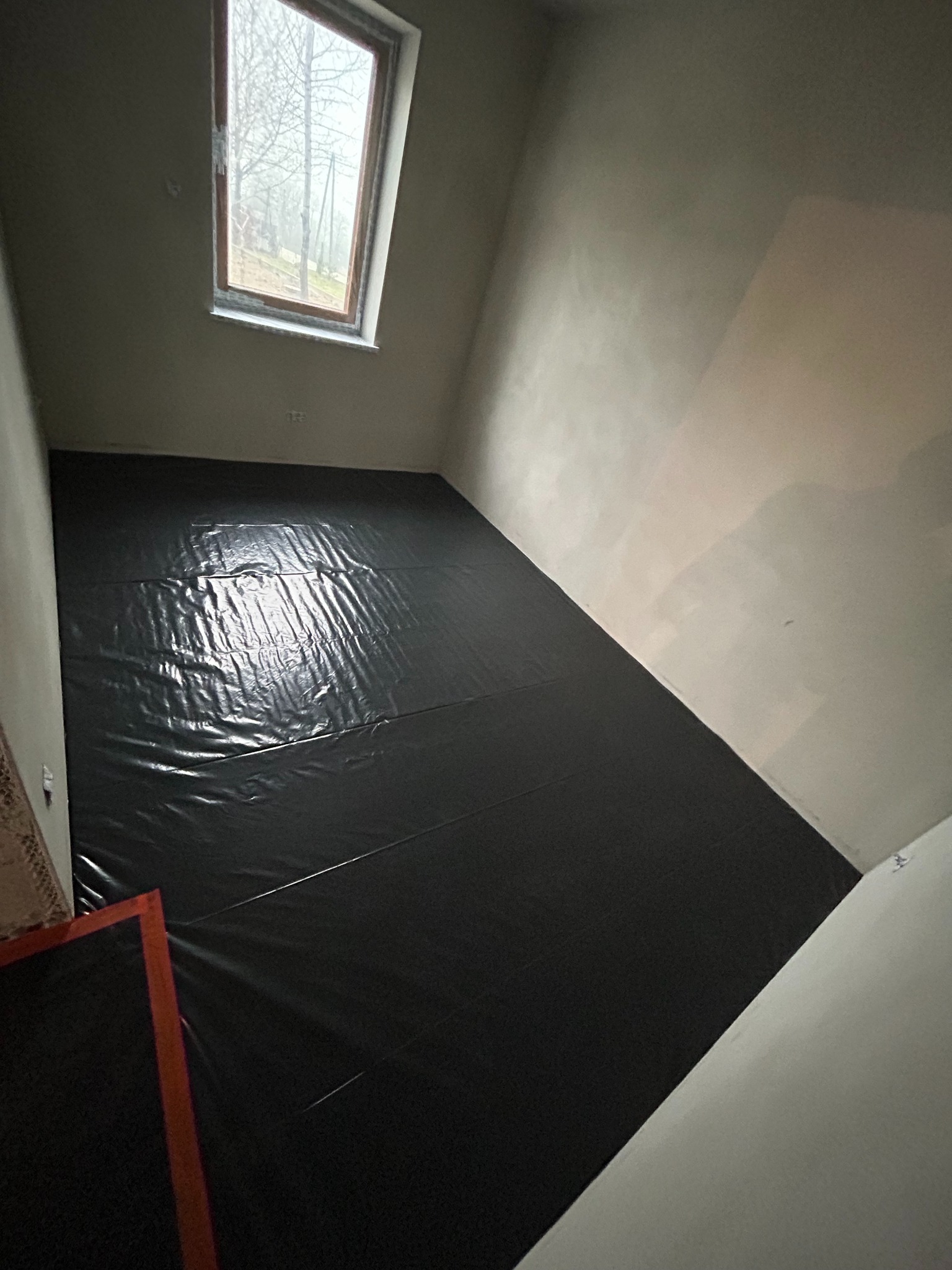 Przygotowanie podłoża: czarna folia na podłodze w surowym pomieszczeniu z oknem. Ściany przygotowane do gładzi. Widoczny fragment grzejnika podłogowego.