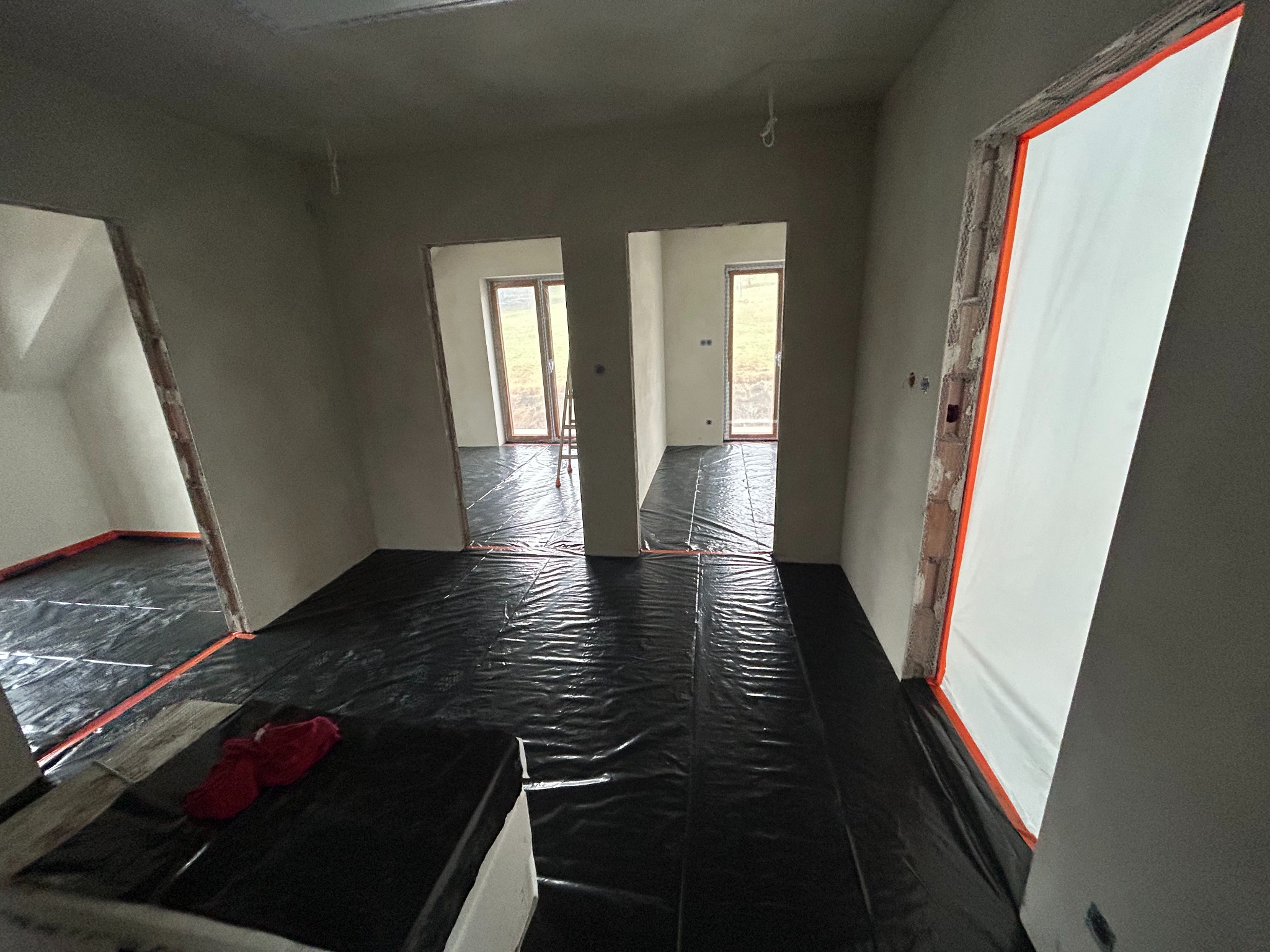 Wnętrze w trakcie prac wykończeniowych: ściany po gładziowaniu, podłoga zabezpieczona folią, widoczne ościeżnice drzwiowe z folią ochronną.